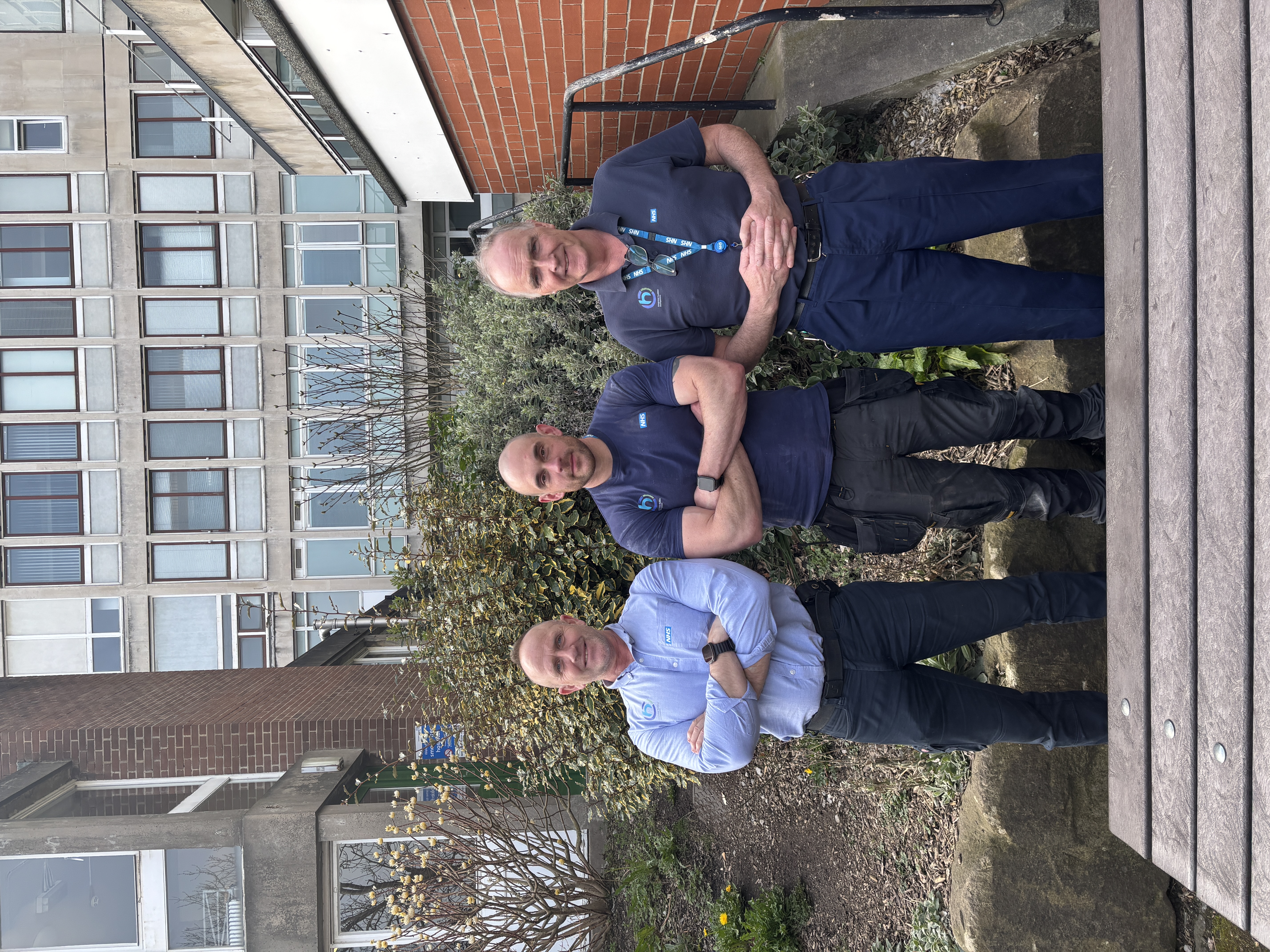 Three smiling men with arms crossed stood in front of a hospital building