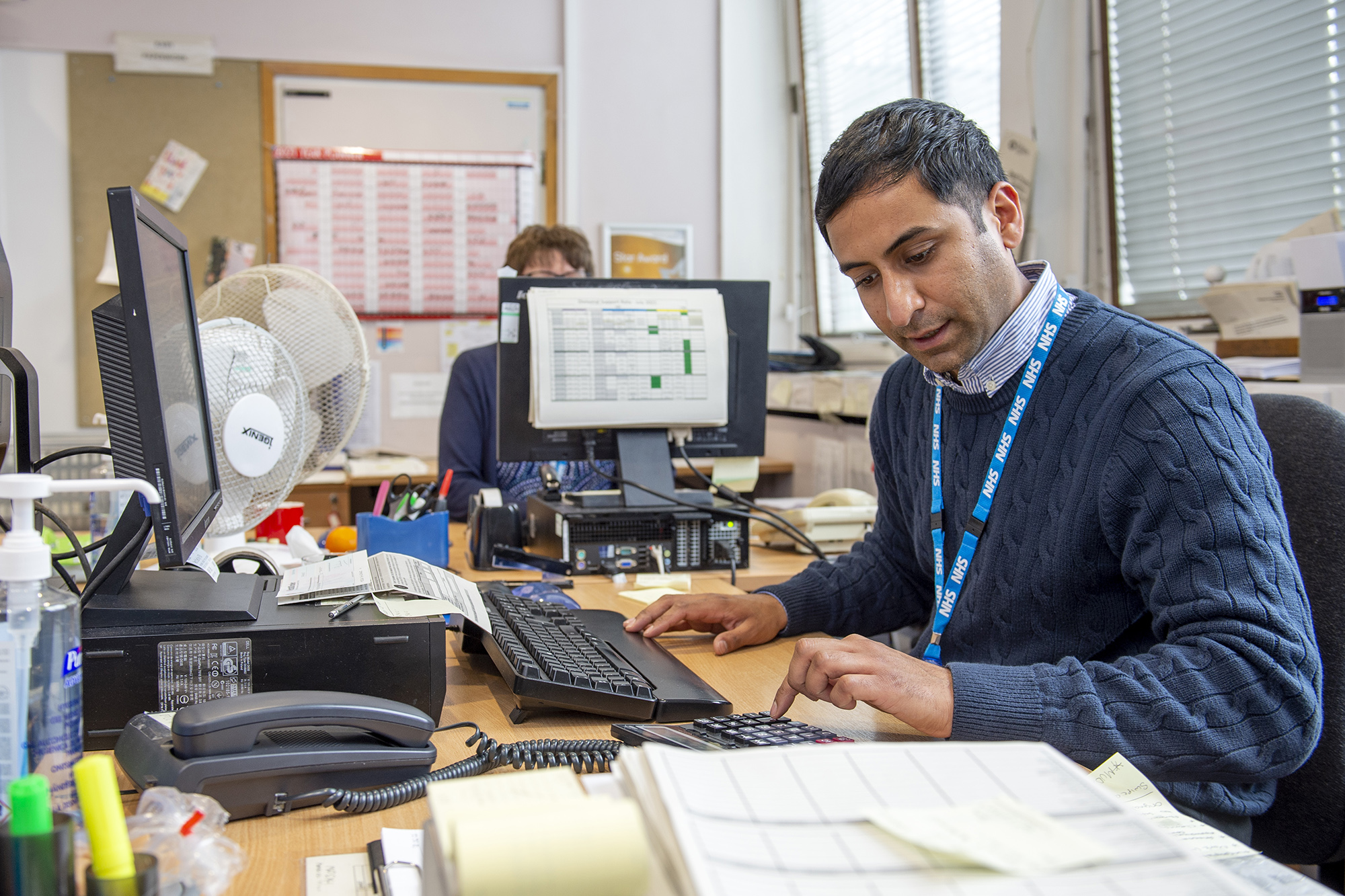 Male member of staff working at his desk which has PC's on using a calculator.