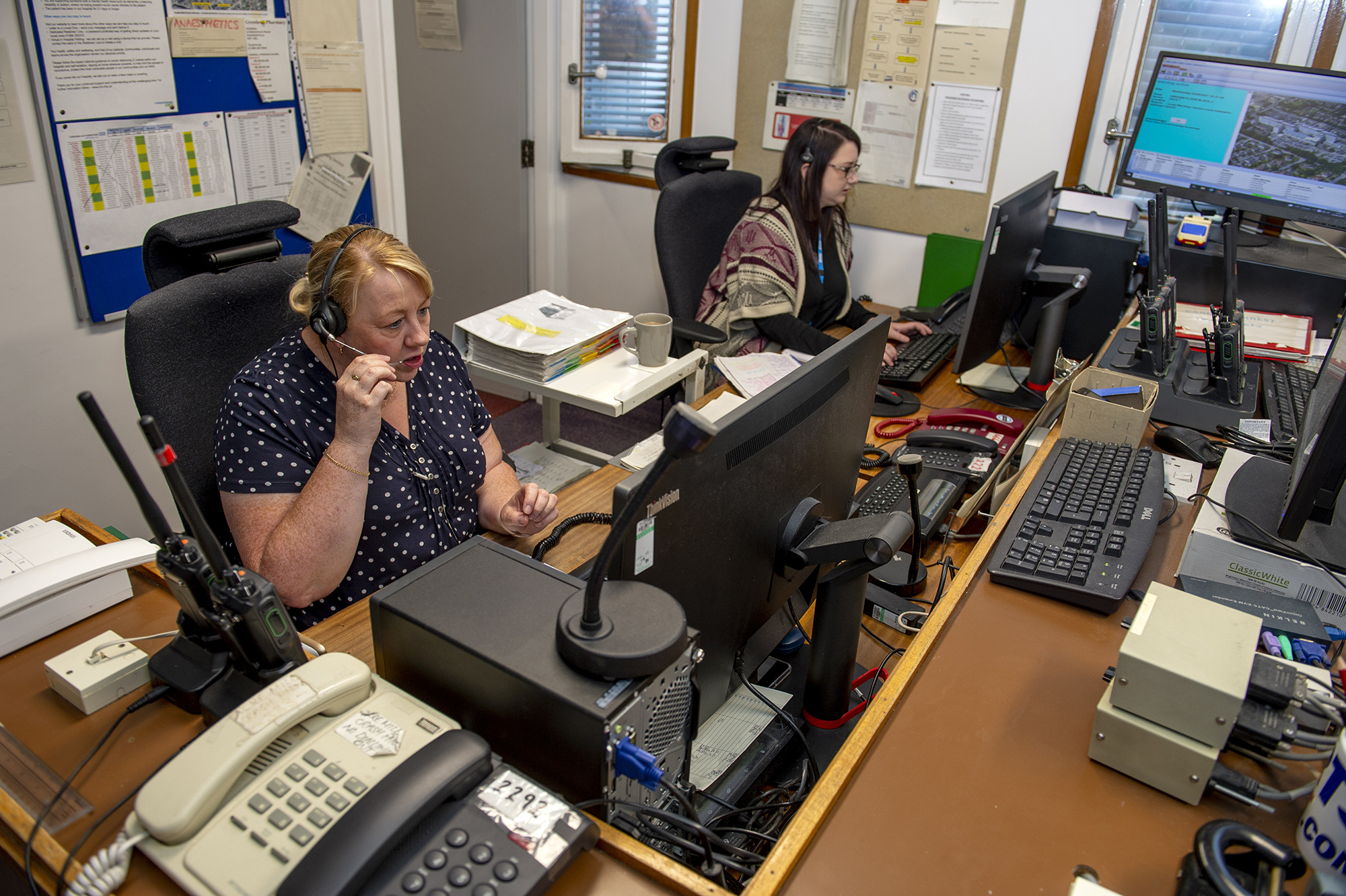 Two female colleagues in a room with PC's with one talking through her headset.
