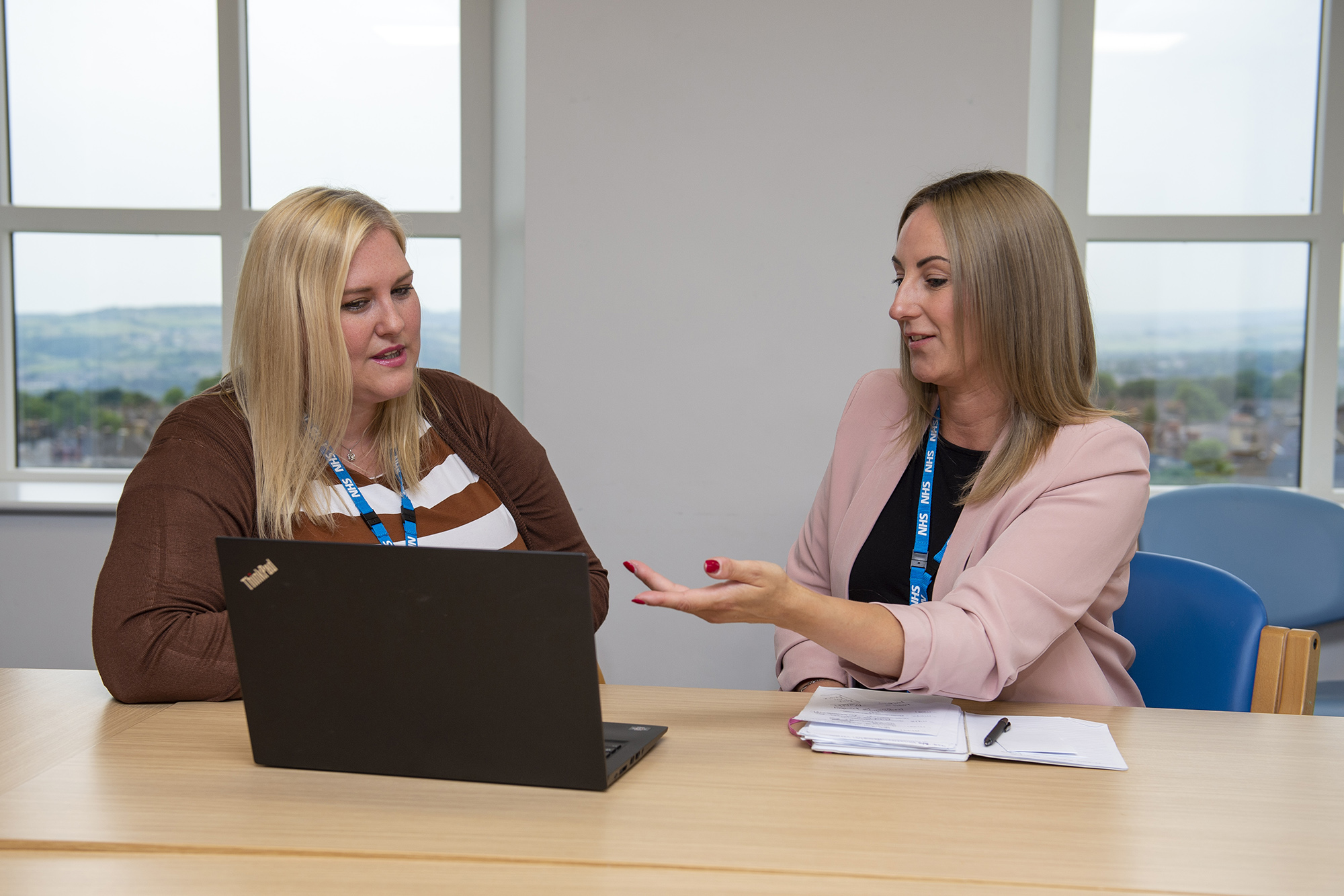 Two female colleagues talking to each other at a desk with a laptop in front of them.